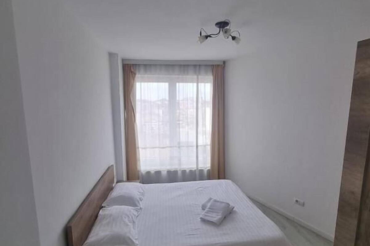 A small, clean room with a single bed, a bedside table, a window with curtains, and a ceiling light fixture.
