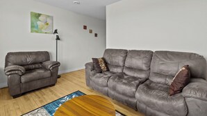 Una sala de estar con dos sofás grises, una lámpara de pie, una mesa redonda de madera y un cuadro en la pared.
