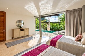 Une chambre avec une grande fenêtre, un lit recouvert d’une courtepointe rouge et beige, une armoire en bois et un fauteuil suspendu près de la fenêtre.