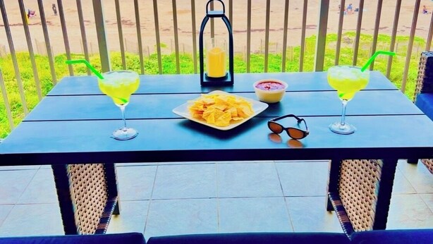 Een tafel met een bord chips, twee drankjes, een lantaarn en zonnebril op een balkon met uitzicht op het strand.