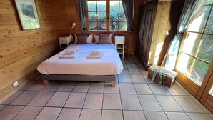 Un dormitorio de cabaña de madera con cama, mesitas de noche, una ventana con cortinas y vistas al verdor exterior.
