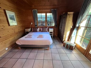 Ein Schlafzimmer in einer Holzhütte mit Bett, Nachttischen, einem Fenster mit Vorhängen und Blick ins Grüne.