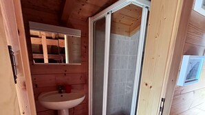 Interior de una cabaña de madera con piso de madera, un lavamanos blanco, un box de ducha y una puerta con picaporte.