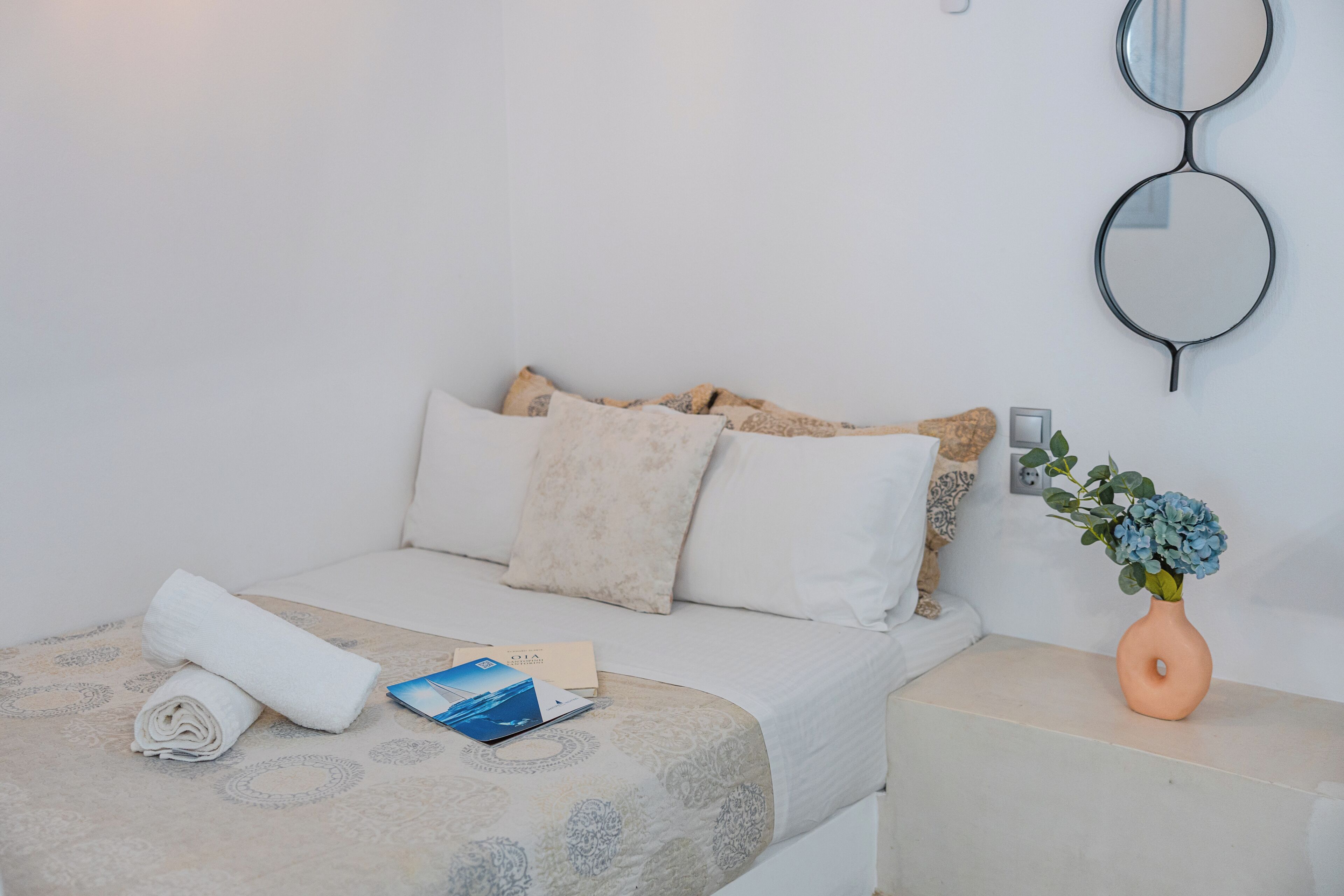 A neatly made bed with pillows, a rolled towel, a book, and a vase with flowers on a bedside table.