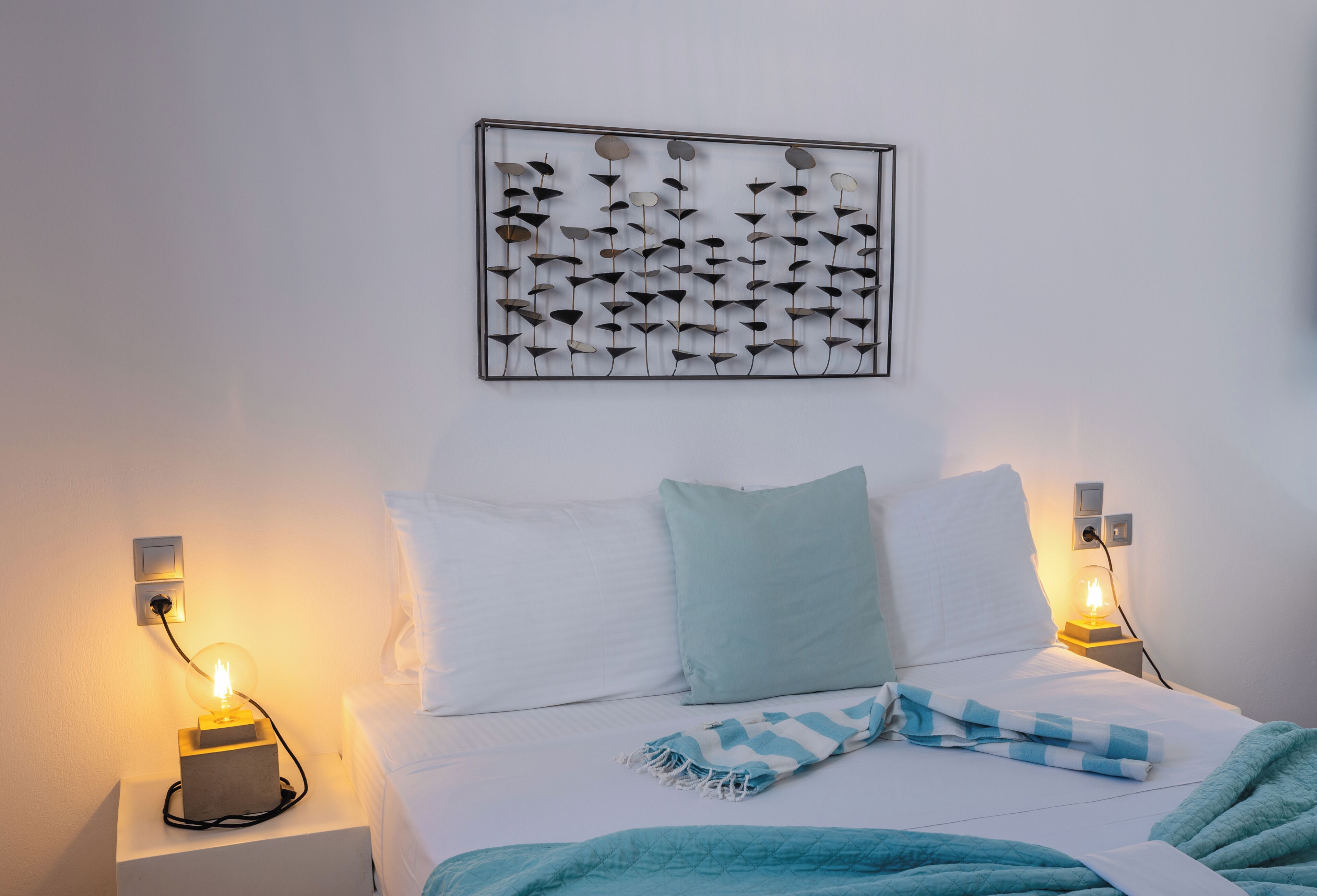 A neatly made bed with white and light blue bedding, a wall-mounted abstract art piece, and bedside lamps on either side.
