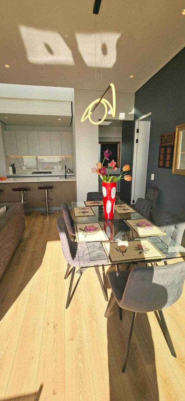 Ein moderner Essbereich mit einem Glastisch, grau gepolsterten Stühlen und einer roten Vase mit Blumen.