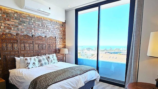 A hotel room with a large bed, a brick wall, and a view of the city and sea. - Luxury modern 3 bed Penthouse apartment in Green Point near V&A Waterfront, CBD (Cape Town)