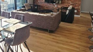 A modern living room with a brick wall, a flat-screen TV, and a dining area with a glass table and chairs. - Luxury modern 3 bed Penthouse apartment in Green Point near V&A Waterfront, CBD (Cape Town)