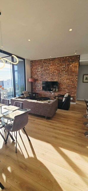A modern living room with a brick wall, a flat-screen TV, and a dining area with a glass table and chairs. - Luxury modern 3 bed Penthouse apartment in Green Point near V&A Waterfront, CBD (Cape Town)