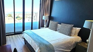 A hotel room with a large bed, a nightstand, a lamp, an air conditioning unit, and a view of a pool and buildings. - Luxury modern 3 bed Penthouse apartment in Green Point near V&A Waterfront, CBD (Cape Town)