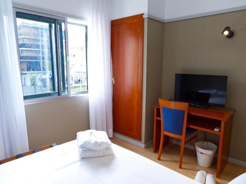 Een hotelkamer met een bed, een televisie op een houten standaard, een stoel, een raam met gordijnen en een deur.