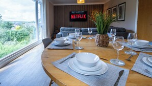 Ruokailutila, jossa on neljälle katettu ruokapöytä, televisio, jossa näkyy Netflix, ja näkymä vehreään maisemaan.