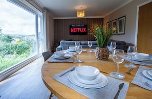 Ein Essbereich mit Tisch für vier Personen, einem Fernseher, der 'NETFLIX' zeigt, und Blick ins Grüne.