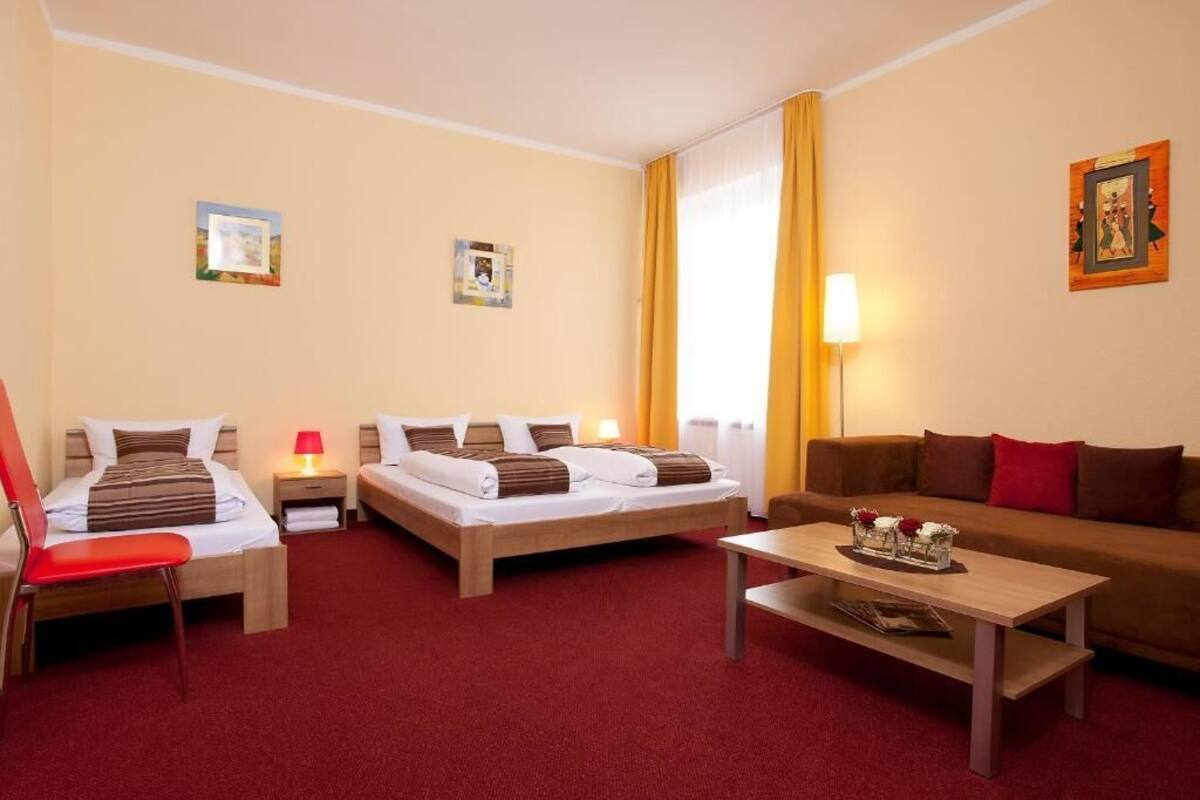 A hotel room with two single beds, a sofa, a red chair, a wooden table, and a lamp.