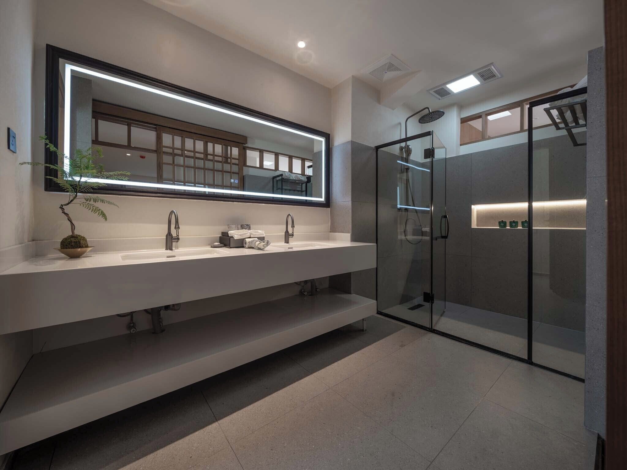 A modern bathroom with a large mirror, two sinks, a glass shower enclosure, and a potted plant.