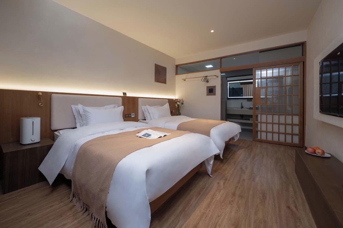 A hotel room with two beds, a wooden floor, a bedside table with a lamp, and a sliding door leading to a bathroom.