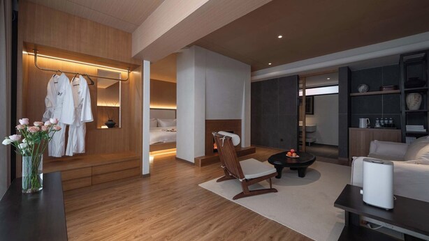 A modern hotel room with a wooden floor, a bed, a seating area with a table, a small fireplace, and a wardrobe with hanging robes. - Shangri-La City Yunjian Songyue (Deqin)