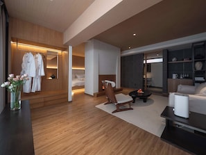 A modern hotel room with a wooden floor, a bed, a seating area with a table, a small fireplace, and a wardrobe with hanging robes. - Shangri-La City Yunjian Songyue (Deqin)