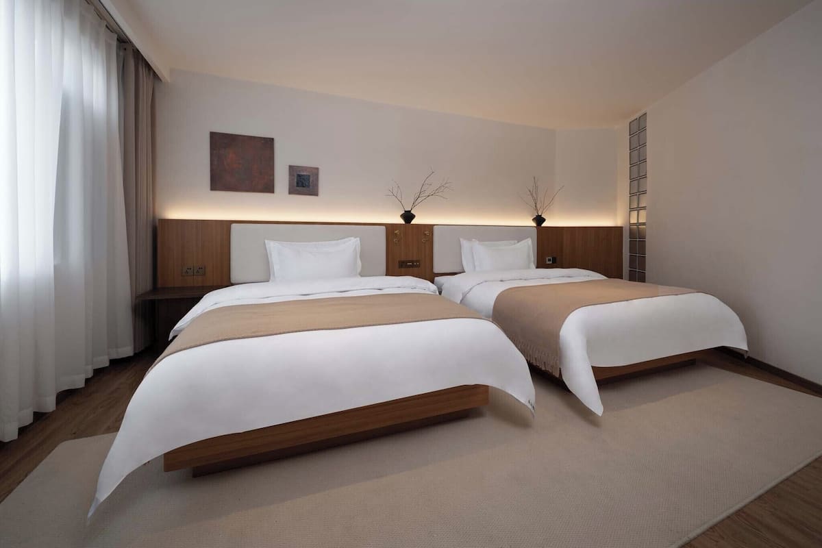 A hotel room with two beds, a nightstand, a wall-mounted light, and a window with curtains.