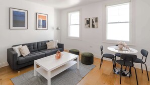 En moderne stue med en grå sofa, et hvidt sofabord og grønne skamler. Væggene er prydet med indrammede kunstværker, og der er et rundt bord med stole.