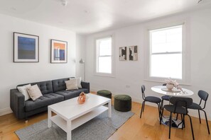 Ein modernes Wohnzimmer mit einem grauen Sofa, einem weißen Couchtisch und grünen Hocker. An den Wänden befinden sich gerahmte Kunstwerke und es gibt einen runden Tisch mit Stühlen.