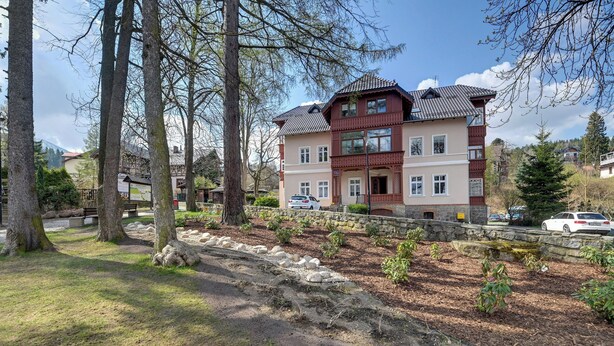 Un edificio a più piani con facciata in pietra, vialetto lastricato e giardino curato con alberi e arbusti.