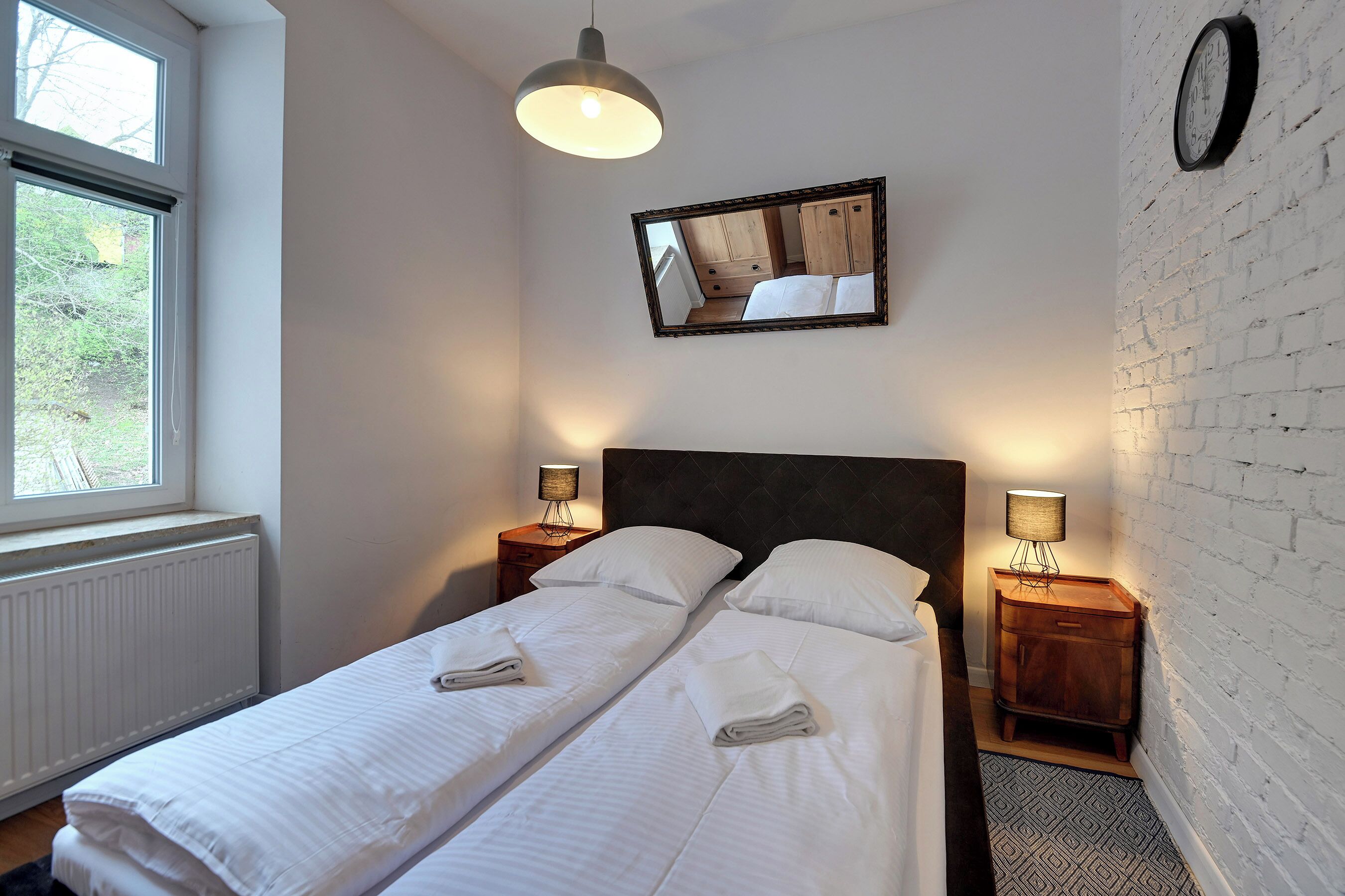 Una cama doble con sábanas blancas, dos mesitas de noche con lámparas, un reloj de pared y una ventana con vistas a zonas verdes.