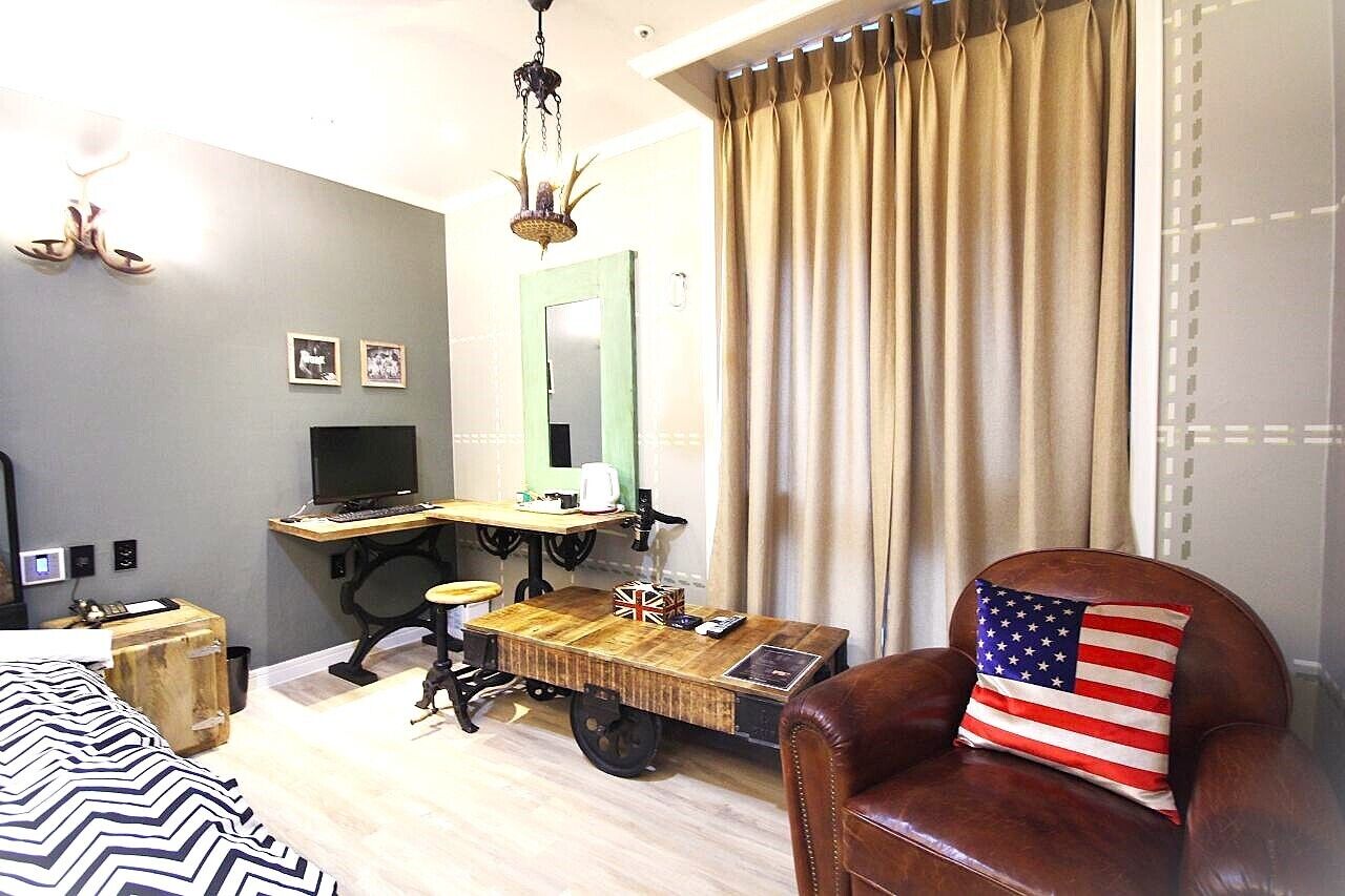 Una stanza con un letto, una scrivania con un computer, una sedia con un cuscino con la bandiera americana e una finestra con tende.
