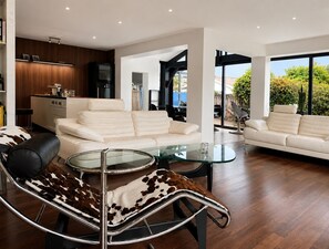 A modern living room with a glass coffee table, leather sofas, and a dining area with a kitchen in the background.
