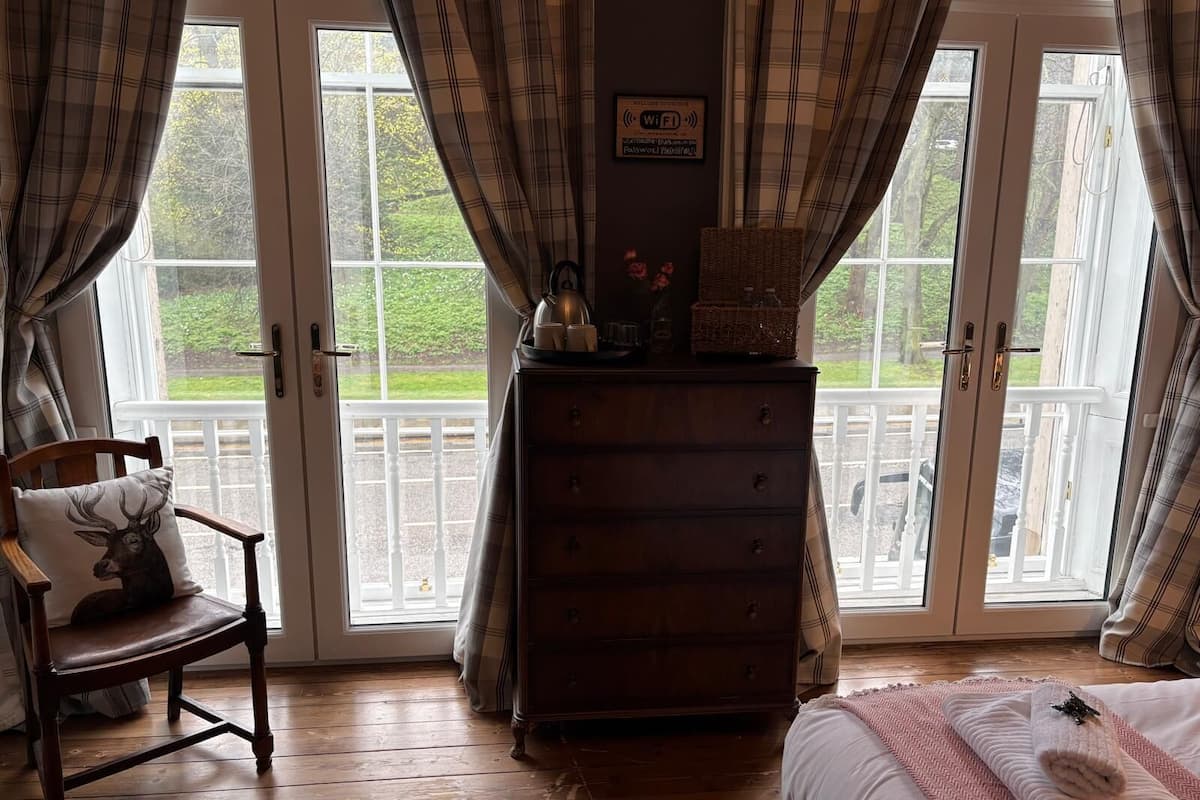 A room with a wooden dresser, a chair with a deer pillow, a bed with a rolled towel, and a balcony with a view.