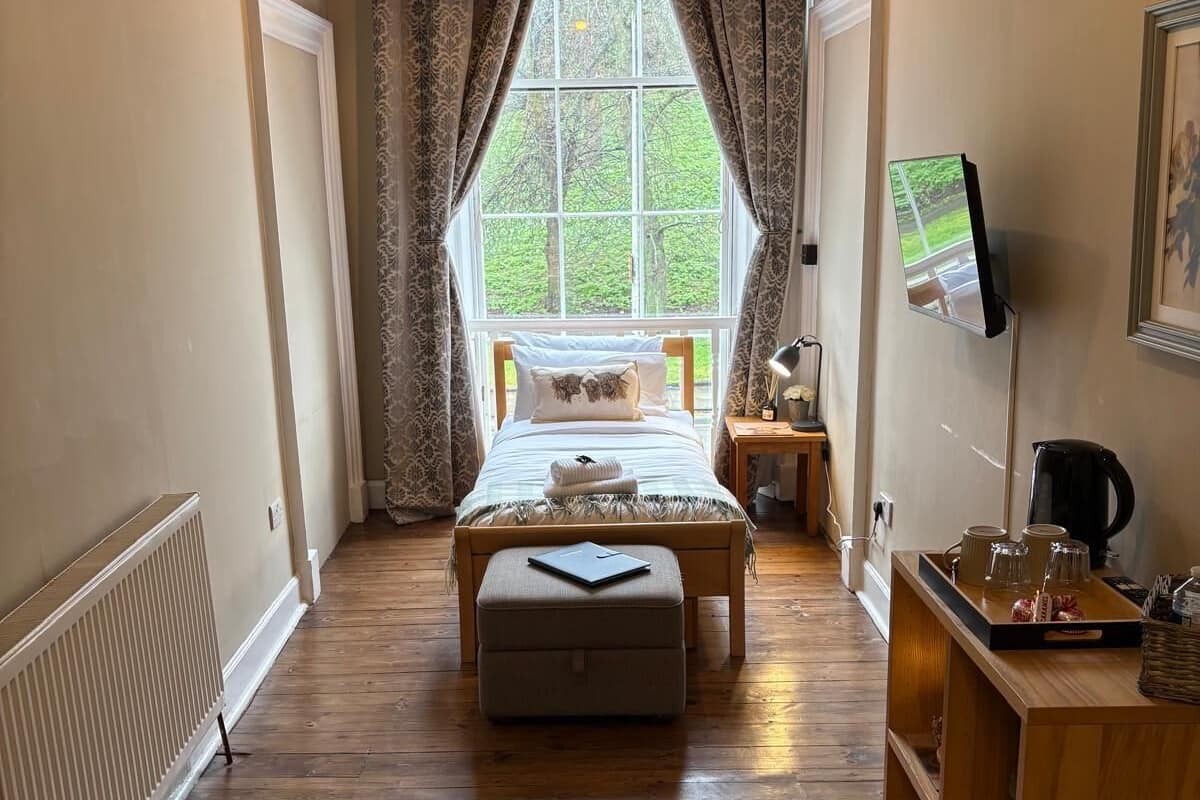 A bedroom with a bed, a bedside table, a TV, a radiator, and a window with curtains.