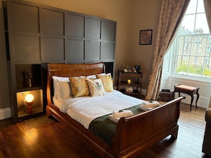 A wooden bed with a headboard, a bedside table with a lamp, and a window with a view of buildings.
