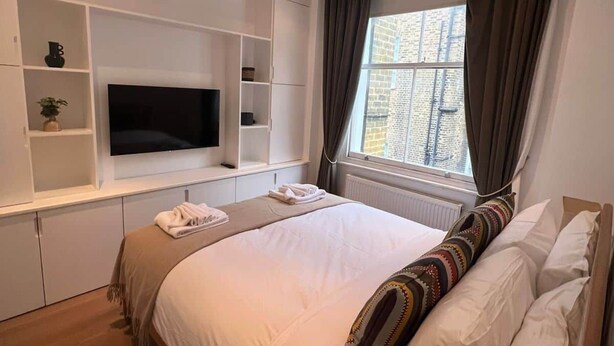 Ett sovrum med en säng, en inbyggd TV, förvaringsmöbler och ett fönster med gardiner.