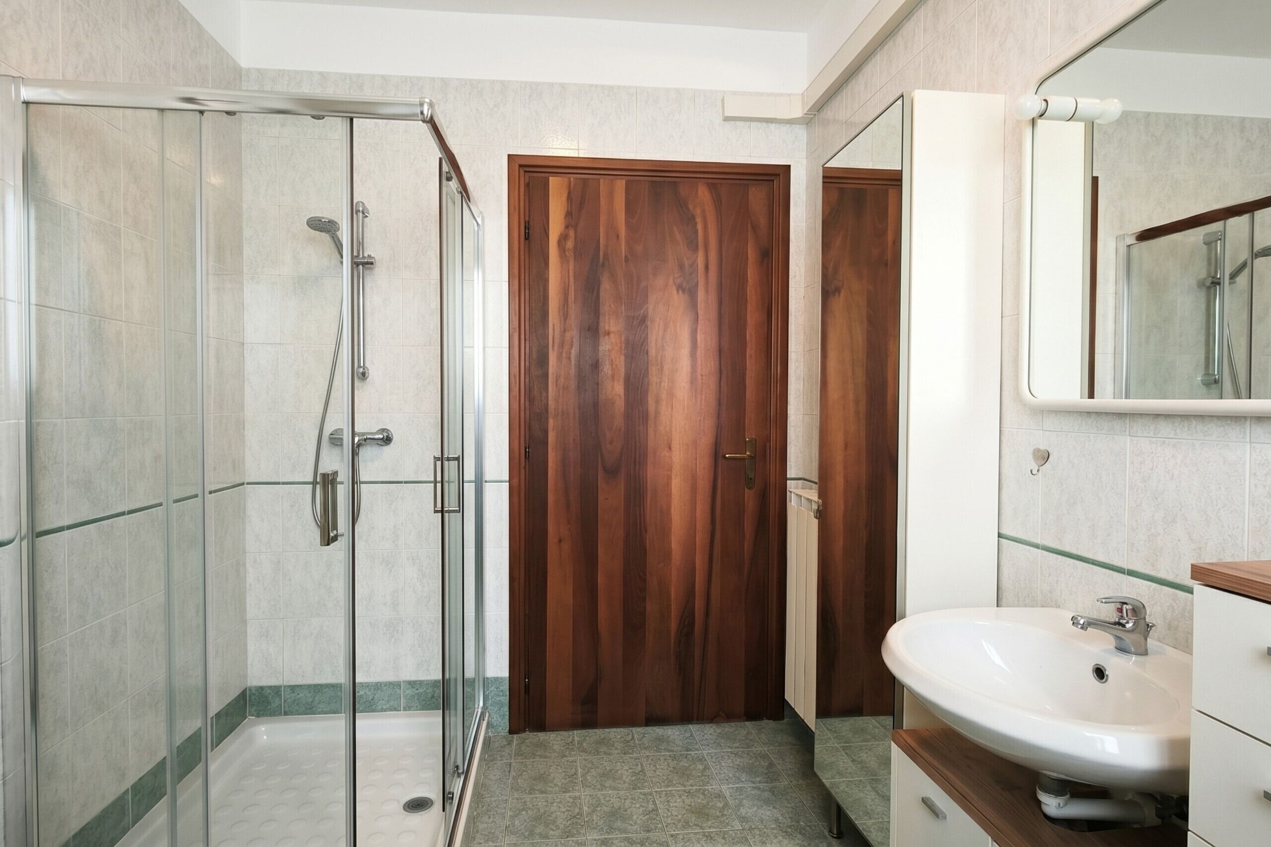 A bathroom with a glass shower enclosure, a wooden door, and a white sink with a mirror.