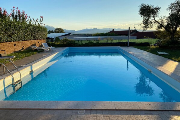 A rectangular swimming pool with clear blue water, surrounded by a paved deck, a brick wall, and a row of white lounge chairs.