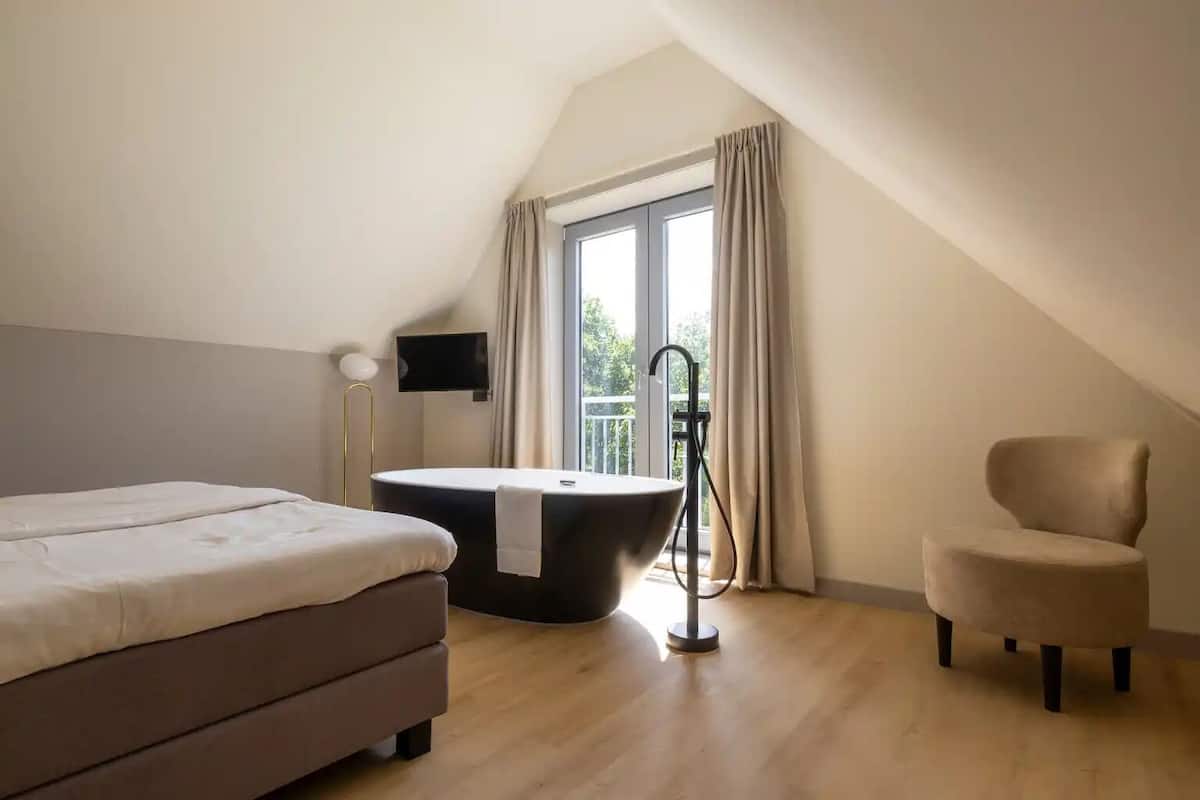 A bedroom with a bed, a bathtub, a chair, a TV, and a window with curtains.