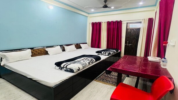 A bedroom with a bed, a table, a chair, a ceiling fan, and a door. - Vandana Palace (Ayodhya)