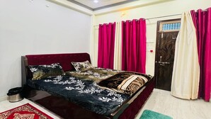A bedroom with a bed, a ceiling fan, a window with curtains, and a door. - Vandana Palace (Ayodhya)