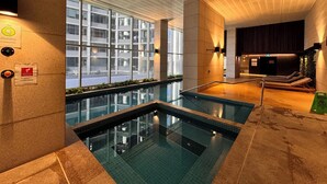 A modern indoor pool area with clear glass walls, a ceiling with a grid pattern, and a view of a building outside. - Luxury Apartment | 2BR City Views | Gym+Pool+Spa+Sauna (Melbourne)