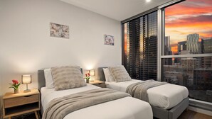 A hotel room with two beds, a nightstand with a lamp, a window with a cityscape view, and artwork on the wall. - Luxury Apartment | 2BR City Views | Gym+Pool+Spa+Sauna (Melbourne)