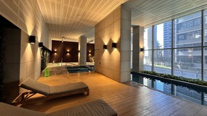 A modern indoor pool area with wooden flooring, lounge chairs, and a view of the city through large windows. - Luxury Apartment | 2BR City Views | Gym+Pool+Spa+Sauna (Melbourne)