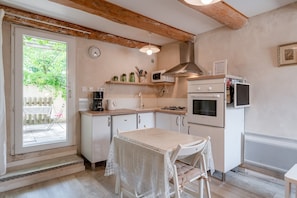 Een keuken met witte kastjes, een tafel met een kantkleed, een klok aan de muur, en een raam met uitzicht op groen.