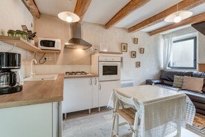 A kitchen with a microwave, stove, and oven, a dining table with chairs, and a sofa in a living area with a window. - Cottage 'Gîte Chez Augustine' with Wi-Fi and Air Conditioning (Sarrians)
