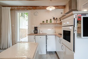 A kitchen with white appliances, a clock, and a window with curtains. - Cottage 'Gîte Chez Augustine' with Wi-Fi and Air Conditioning (Sarrians)