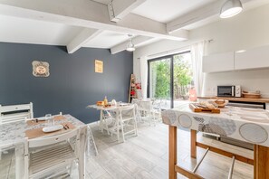Uma cozinha moderna com armários brancos, uma área de refeições com cadeiras e mesa brancas, e uma janela grande que oferece vista para o verde lá fora.