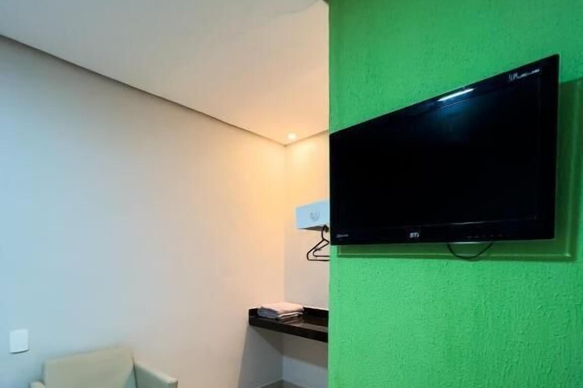 A room with a green wall, a flat-screen TV mounted on it, a small beige chair, and a shelf with a towel.