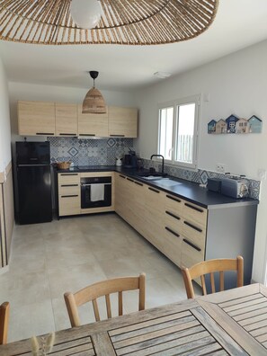 Eine moderne Küche mit Holzschränken, einem schwarzen Kühlschrank, einem Esstisch mit Stühlen und einem großen Fenster.