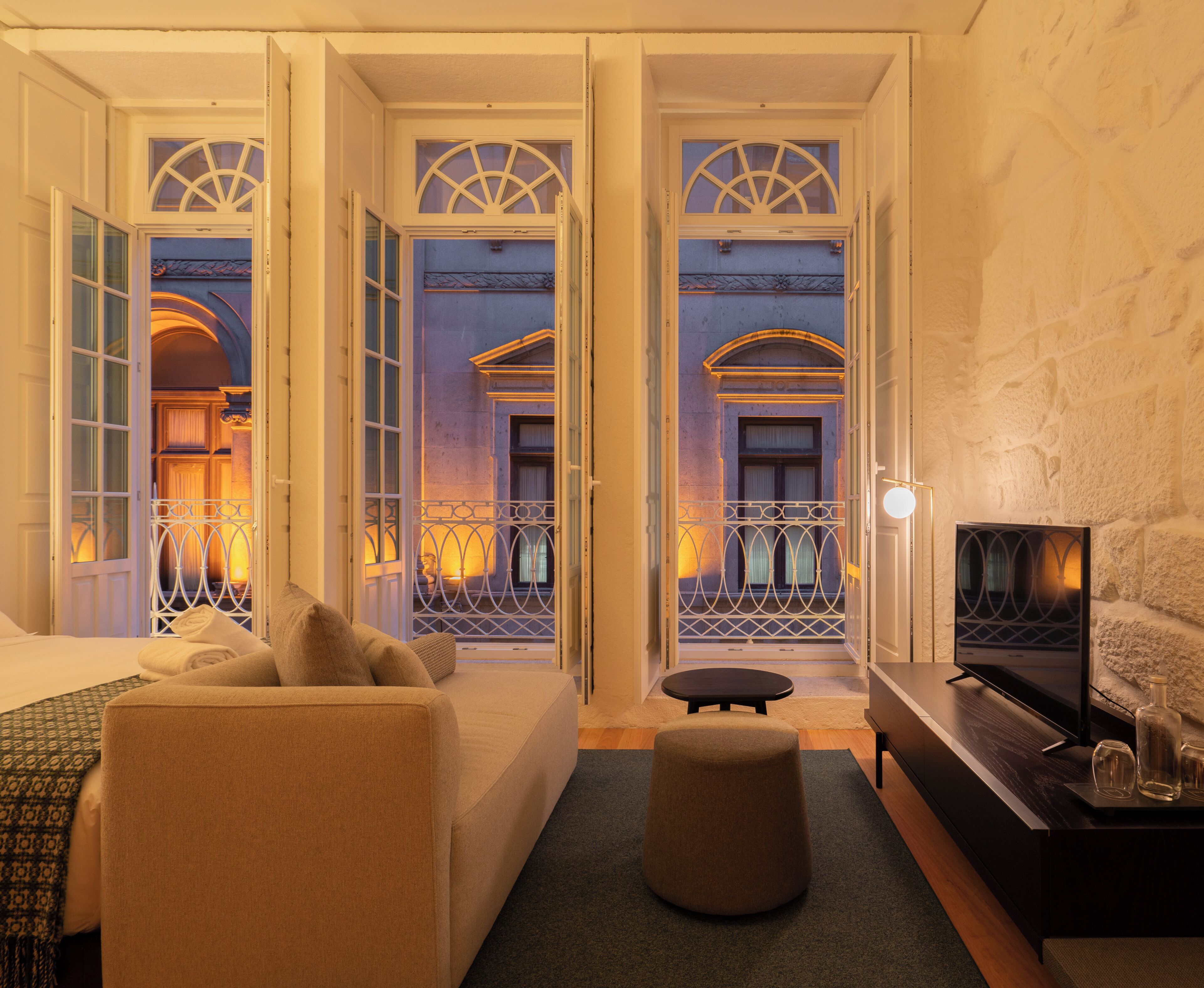 Habitación de hotel con cama, sofá, un pequeño reposapiés, televisión y vistas al balcón exterior.