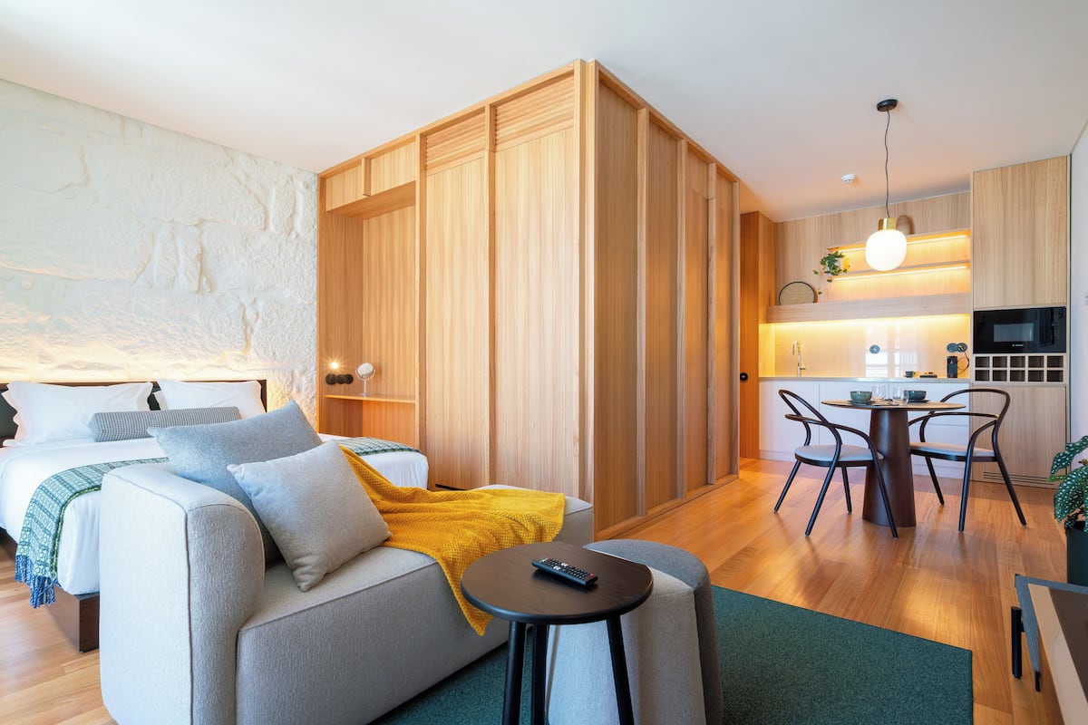 A modern hotel room with a sofa, a small round table, a bed, a wooden wardrobe, a kitchen area with a microwave, and a dining table with chairs.