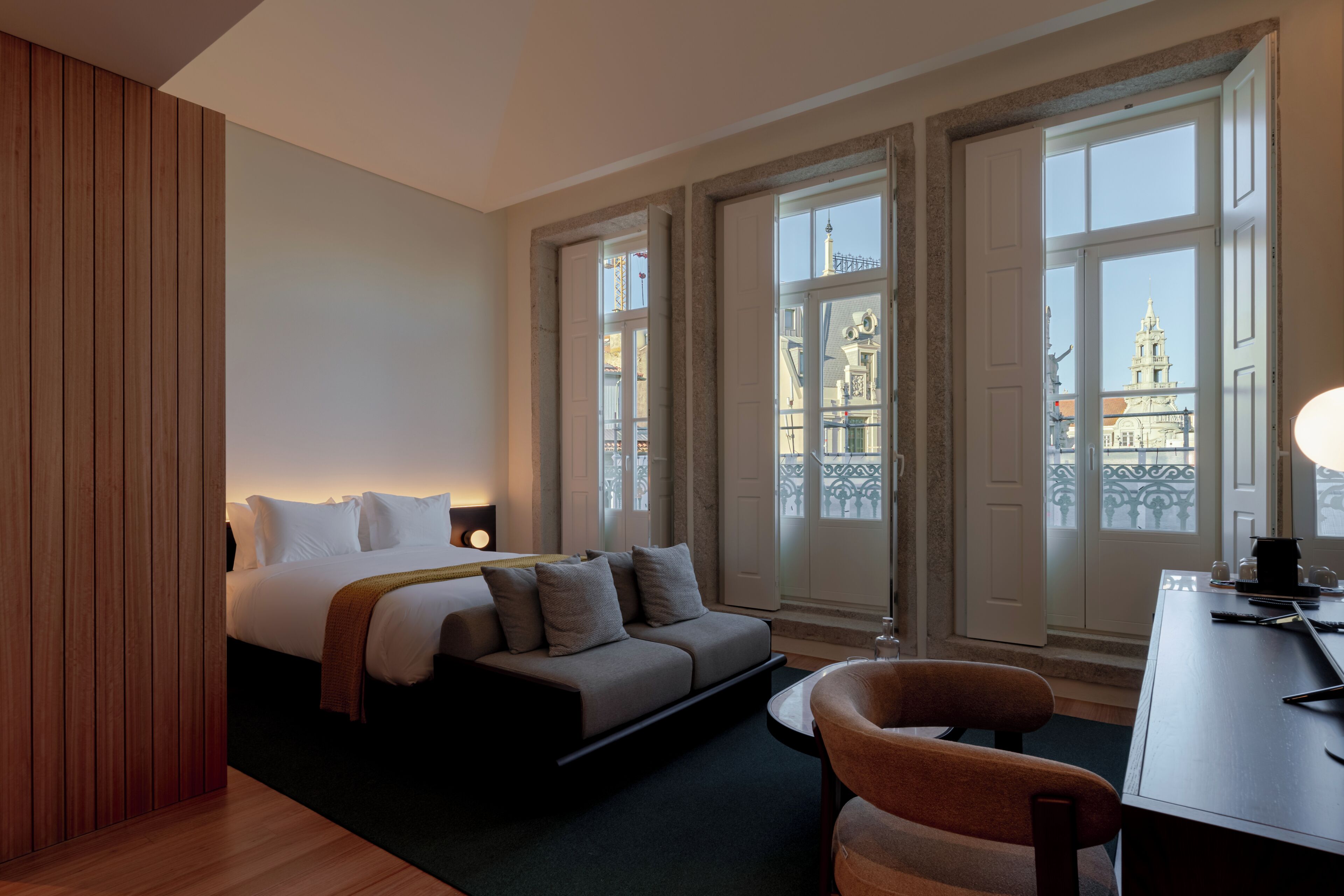 A hotel room with a bed, a desk, a chair, and large windows with a view of buildings.